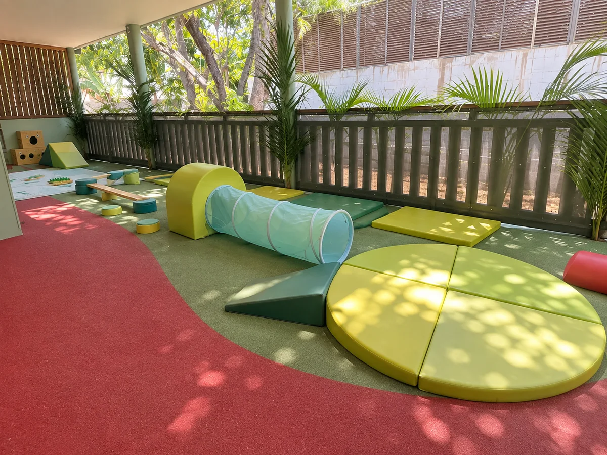 Terrasse ombragée de crèche avec tunnel, modules et tapis de motricité en mousse verts et jaunes sur sol rouge et gazon synthétique, ambiance calme et tropicale, idéale pour présenter les espaces d'éveil.