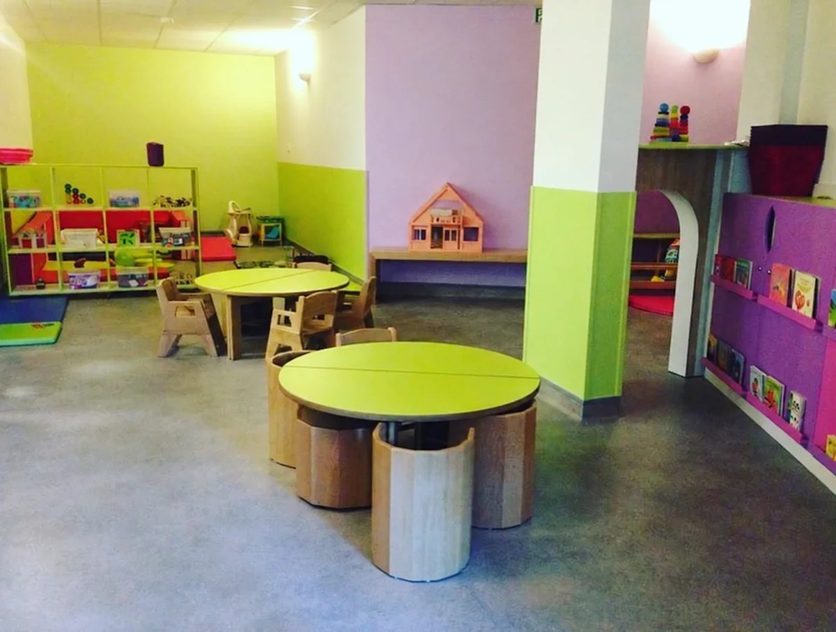 Salle de vie lumineuse d'une crèche aux murs vert anis et mauve, avec tables rondes vertes, tabourets en bois, étagères de jeux et livres, idéale pour présenter les espaces d'accueil.