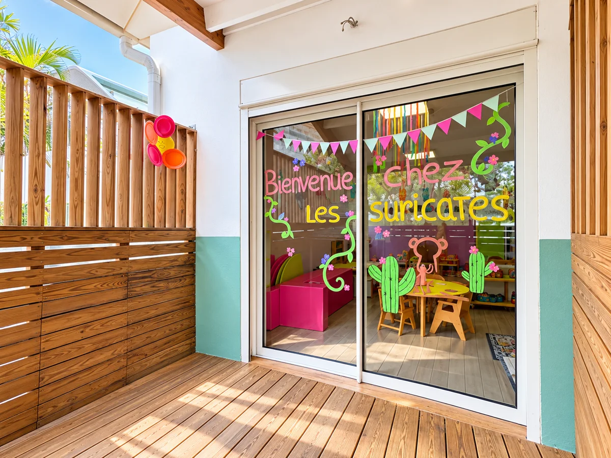 Baie vitrée décorée « Bienvenue chez Les Suricates » avec fanions et cactus colorés, donnant sur une salle de crèche, terrasse en bois ensoleillée ; idéale pour page d'accueil ou présentation de la crèche.